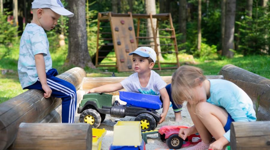 Three children engaged in imaginative play in an outdoor sandbox with toys.