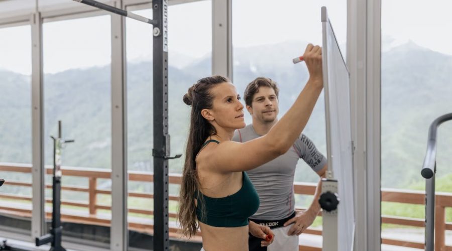 Female fitness coach writes on whiteboard as a male athlete watches in a gym.